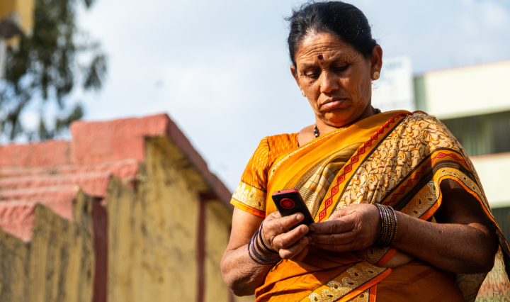 An older Indian woman in a yellow sari stands outside, looking at her phone
