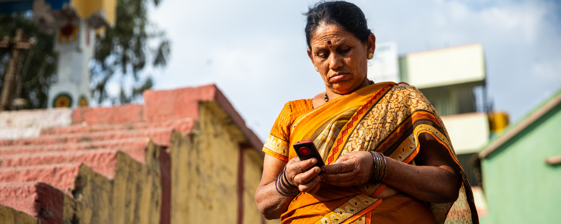 An older Indian woman in a yellow sari stands outside, looking at her phone