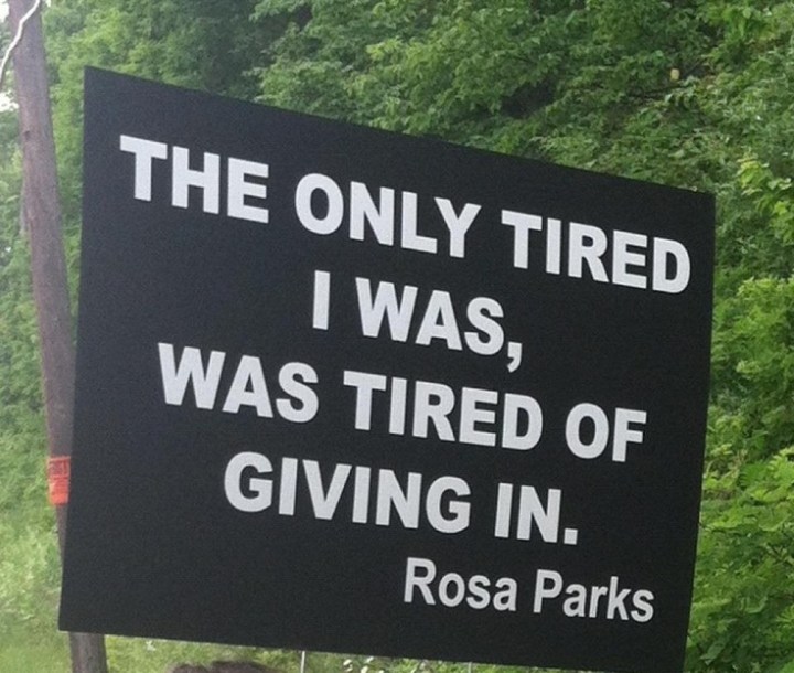 A black sign with white writing, with trees in the background. Text says: The only tired I was, was tired of giving in - Rosa Parks