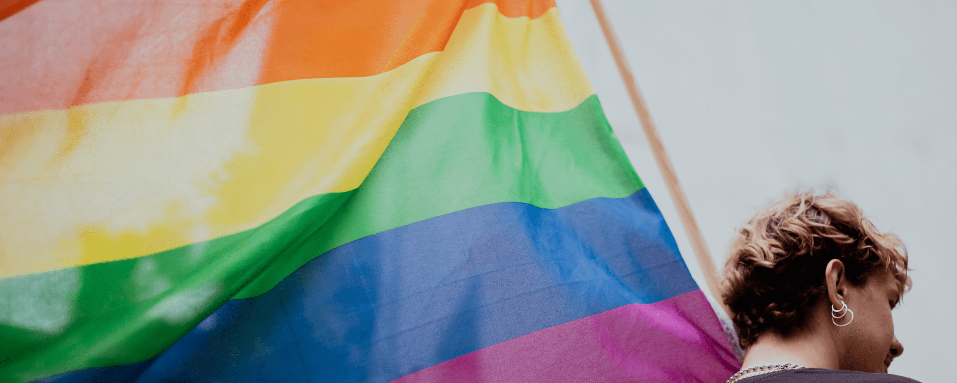 A young person with short strawberry blonde hair and earrings holds a large pride flag in the air