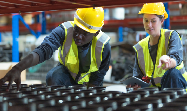 A young Black man touches stock in a warehouse, while a woman points and holds a tablet. They both wear uniforms, hi-vis vests, and yellow hard hats