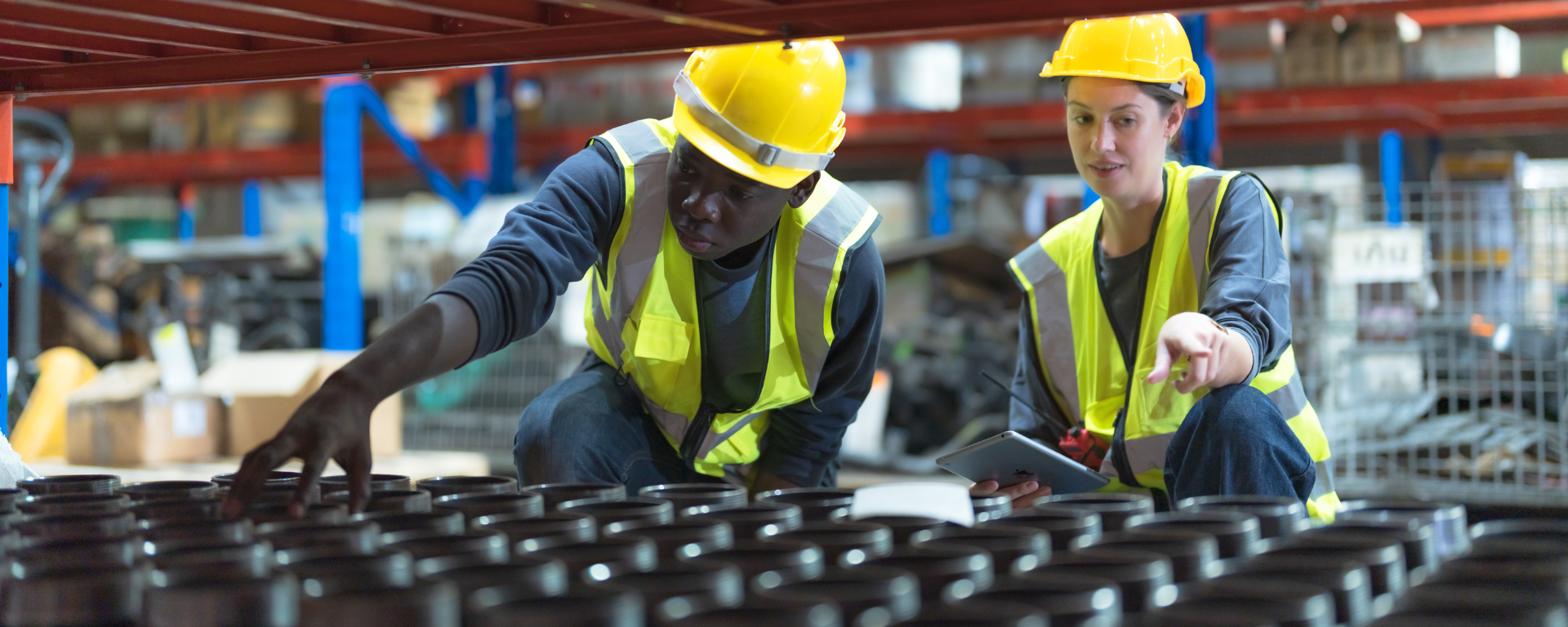 A young Black man touches stock in a warehouse, while a woman points and holds a tablet. They both wear uniforms, hi-vis vests, and yellow hard hats