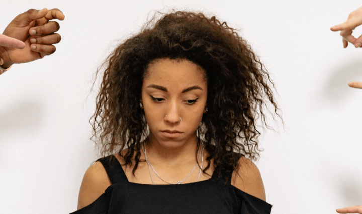 A Black woman stands in the centre looking sad as she glances down, while hands point at her in accusation
