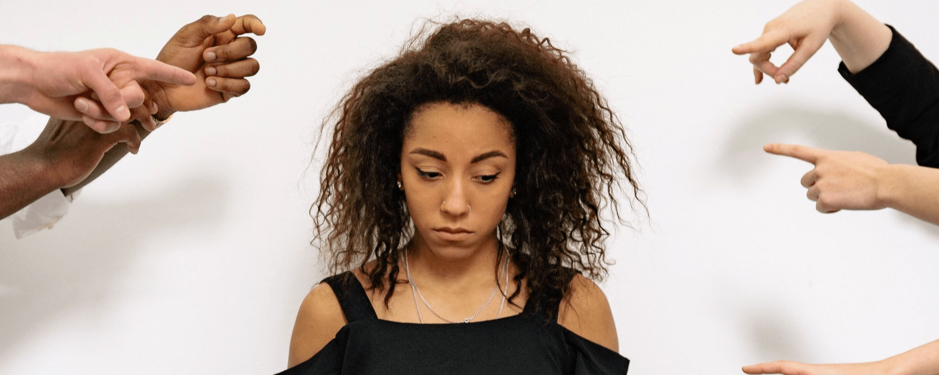 A Black woman stands in the centre looking sad as she glances down, while hands point at her in accusation